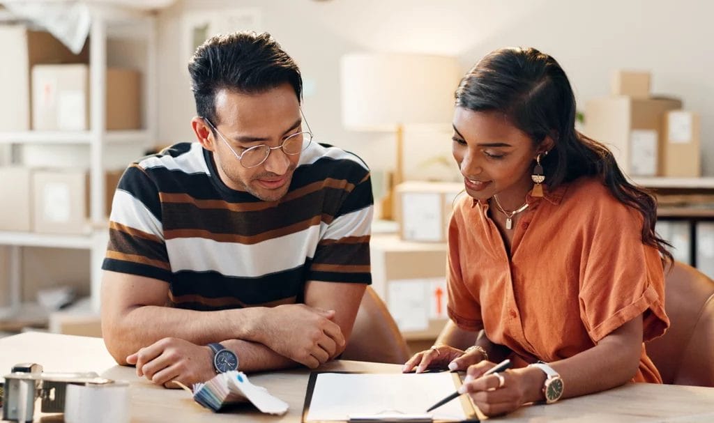 Two small business owners are going over a document on a clipboard in a home office that is full of storage and shipping boxes.