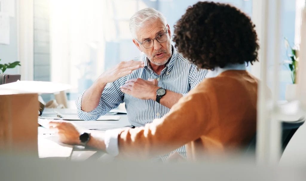 Two business owners are discussing information in a small office space, with one using his hands to help explain a concept to the other person.