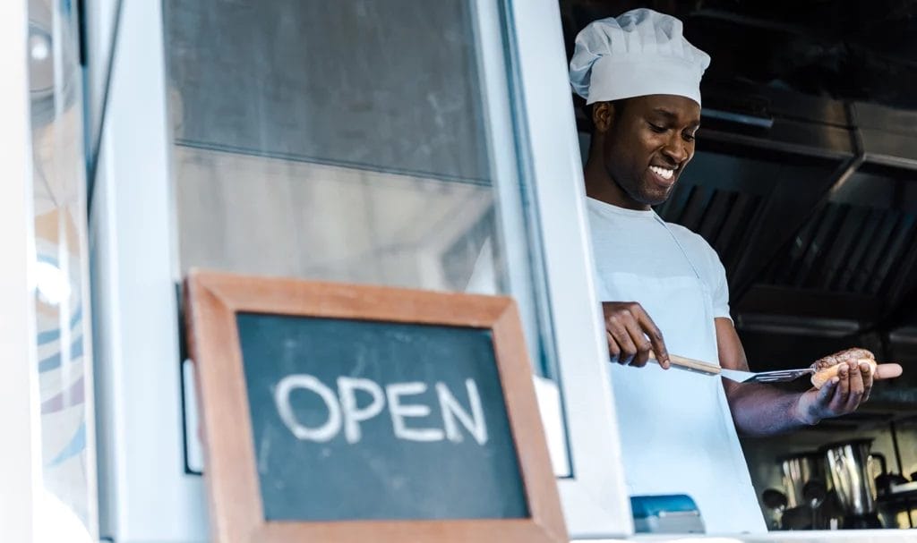 A food truck owner happily prepares a burger inside his truck next to a chalkboard sign on the window that says "Open."
