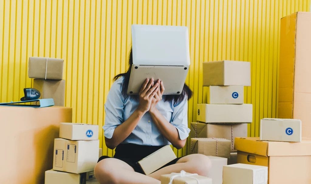 A business owner holds a laptop up to her face while she sits surrounded by product boxes against a yellow background.