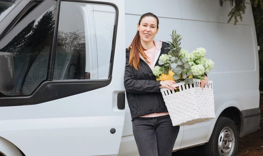 An interior designer smiles at the camera as she gets out of her van holding a basket of fake plants and flowers on her way to set-up for an event.
