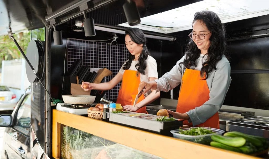 A food truck owner and her employee happily prepare ingredients in the truck for the day ahead.