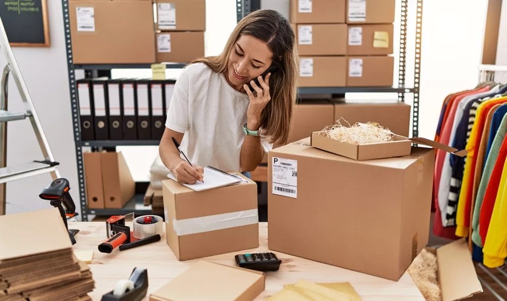 A clothing designer talks on the phone while writing on a notepad in her office, surrounded by boxes, clothing samples, and shipping materials.