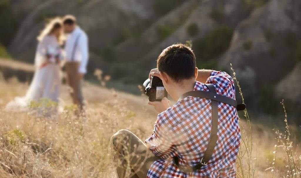 A photographer kneels in a grassy field to capture the perfect shot of a bride and groom posing in the distance.