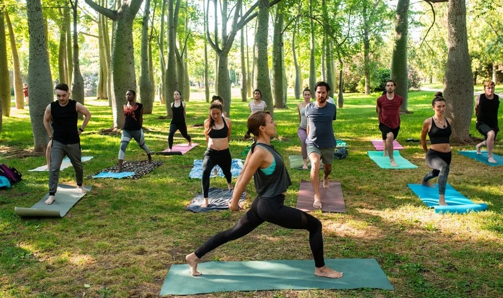 A yoga instructor leads an outdoor class in a public park in a small grove of trees. She stands sideways in front of the class to demonstrate the elongated stance to her students.