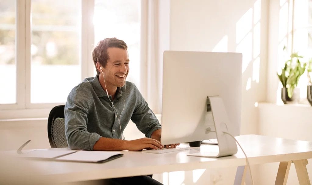 A business owner happily smiles at his computer from his home office while wearing headphones and an open notepad lays beside him on the desk.