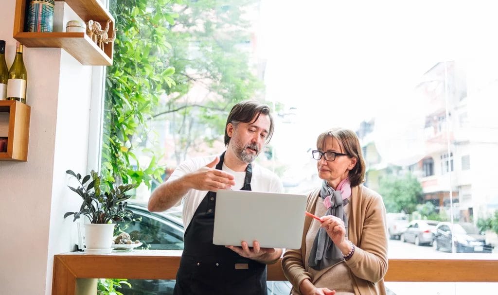 A chef meets with a financial consultant in his cafe as they speak and go over important information on a laptop together.