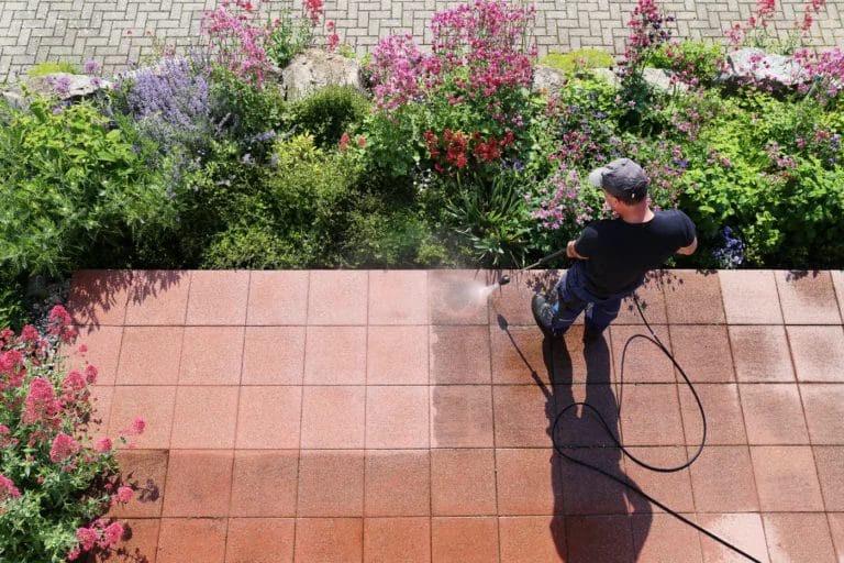 Top view of a man pressure washing a red tile patio surrounded by flowers.