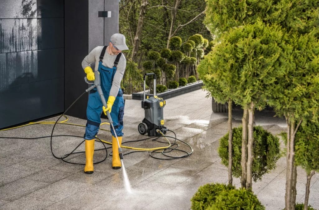 Man in yellow welly boots pressure washing a stone-tile driveway lined with shrubs.