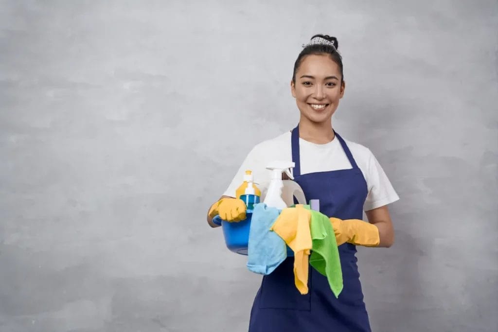 A cleaning professional wearing a white t-shirt, navy apron, and yellow gloves smiles and holds cleaning supplies, including a blue bucket, colorful towels, and spray bottles.