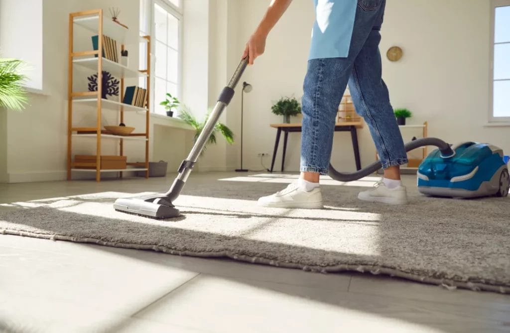 A cleaning professional wearing blue jeans, a blue apron, and white sneakers vacuuming a gray rug in a home with a blue vacuum.