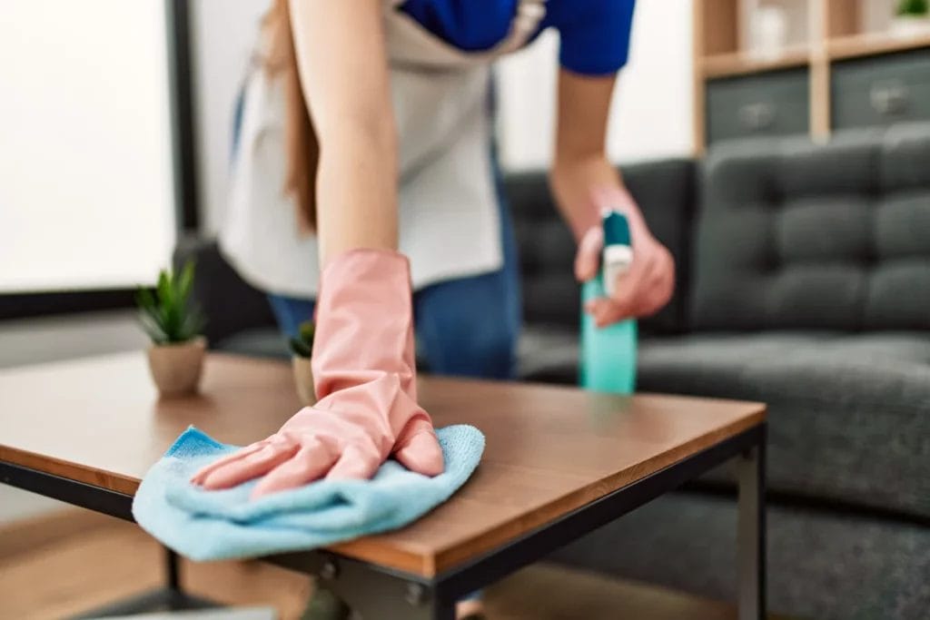 A close-up image of a cleaning professional's pink gloved hands wiping down a wooden coffee table with a blue rag.