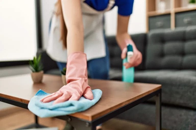 A close-up image of a cleaning professional's pink gloved hands wiping down a wooden coffee table with a blue rag.
