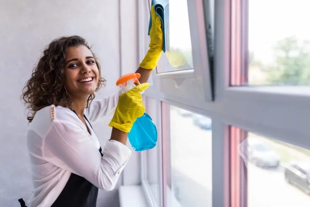 A cleaning professional wearing a white top, black apron, and yellow gloves smiles and cleans a window using blue cleaning solution in a spray bottle and a blue towel.