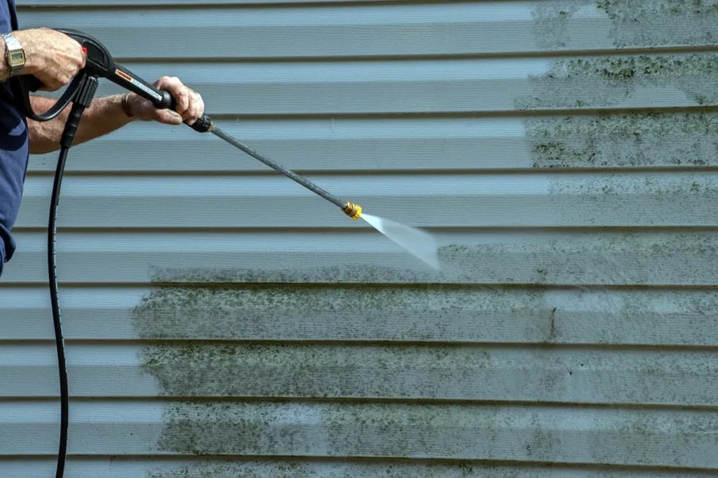 A person pressure washing the side of a home to remove mildew and moss growth.