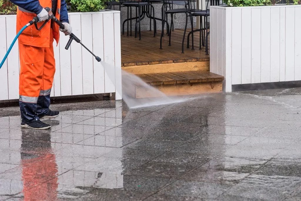 A person wearing bright orange coveralls pressure-washes concrete tiles.