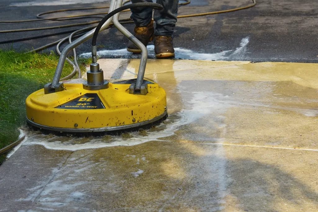 A person uses a round, yellow surface-cleaning pressure washer to clean concrete.