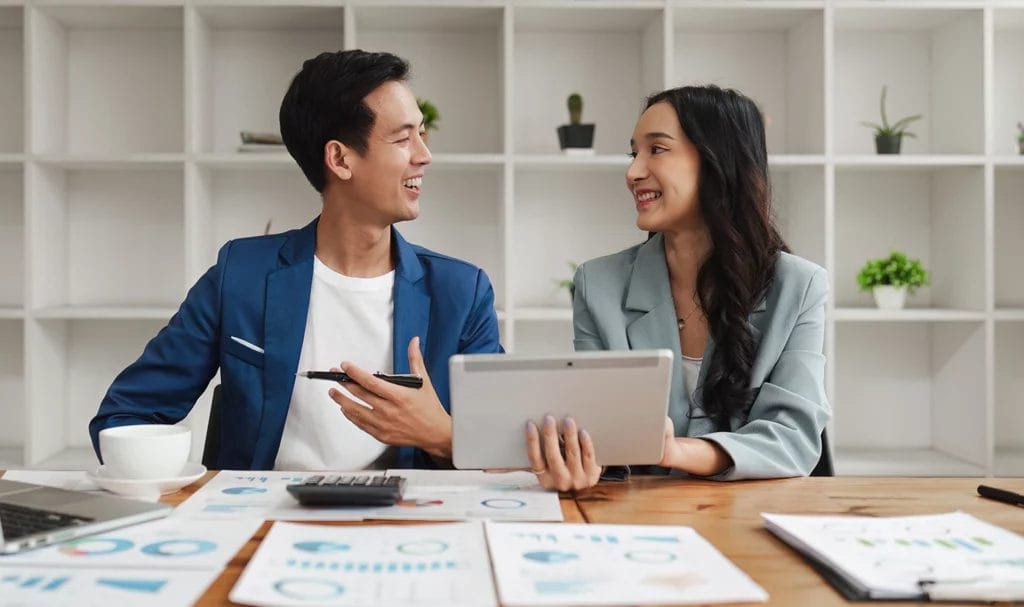 Two business owners consult in a home office with a table and paperwork.