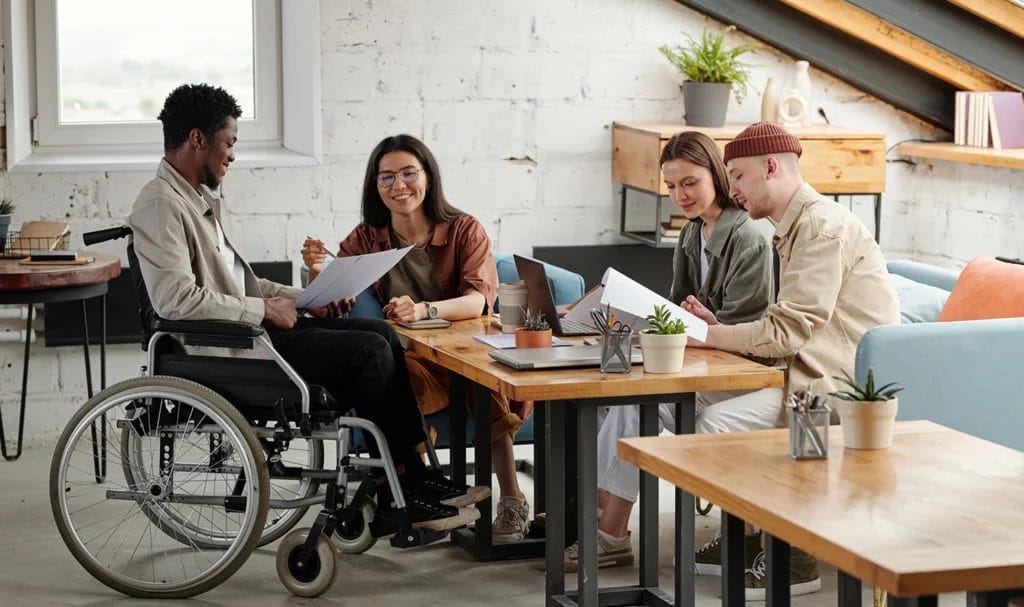 Employees working in a small loft officespace while female economist consultant points at a document held by male colleague in wheelchair.