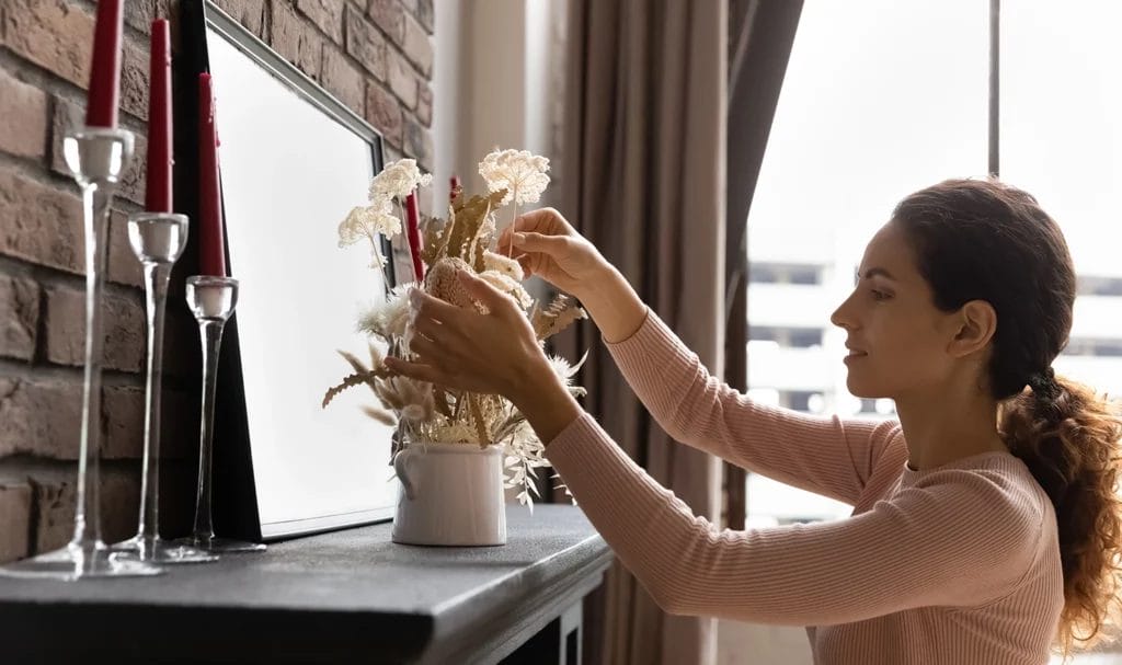 Interior designer decorating a fireplace mantel with dried flowers in vase.