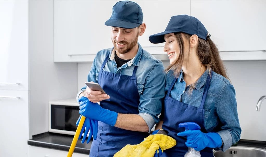 Two professional cleaners in blue apron uniforms standing together with phone as they clean an office kitchen.