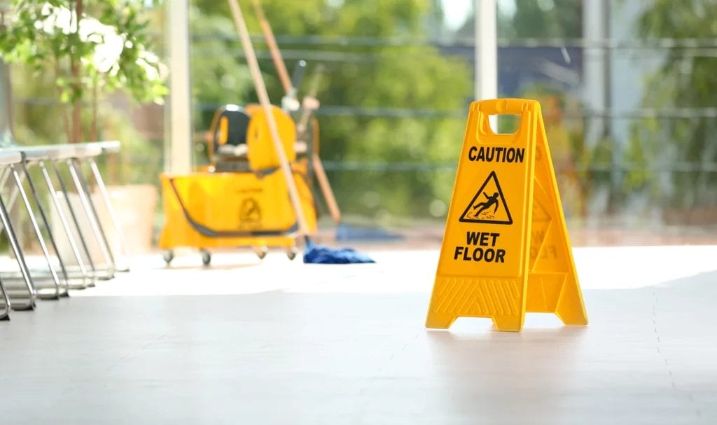 Safety sign with phrase Caution wet floor and blurred mop bucket in background.