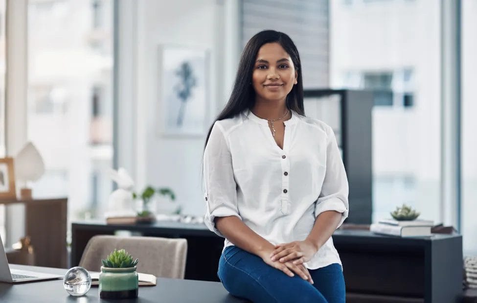 A business consultant wearing a white top and blue jeans smiles and leans against a desk in an office with green plant accents and large windows in the background.