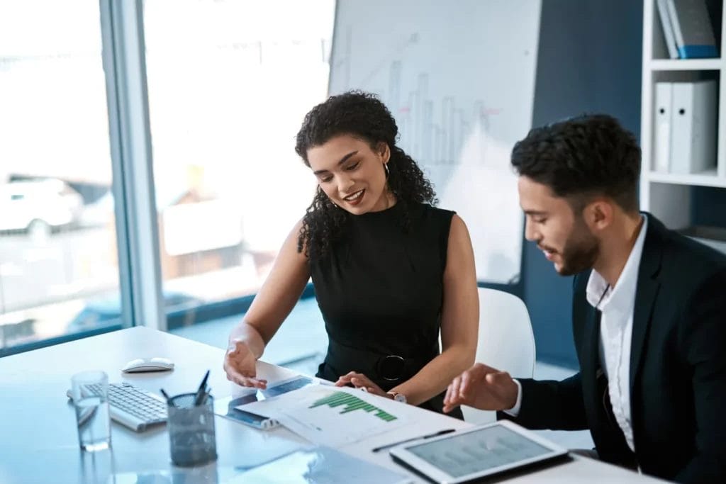 A business consultant wearing a black sleeveless dress reviews a business plan with a client at a white table in an office setting.