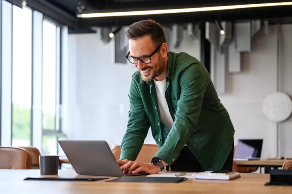 A business consultant wearing black glasses and a green button-up shirt smiles while typing on his laptop at a wooden table in a modern office setting.