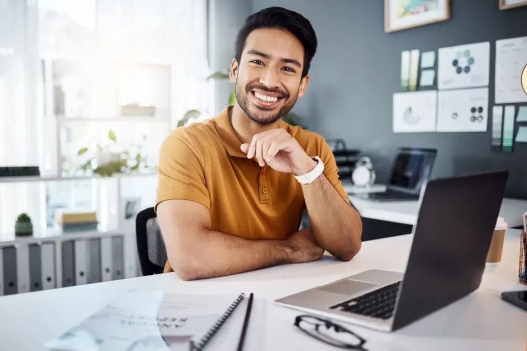 A business consultant wearing a mustard-colored polo shirt smiles while sitting at a white desk with a laptop, business plan, and glasses on it.