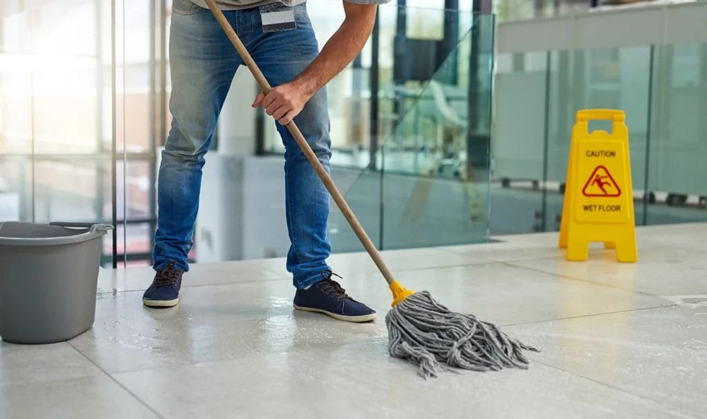 Close-up of a janitor mopping the floors of a building with a caution wet floor sign behind them.