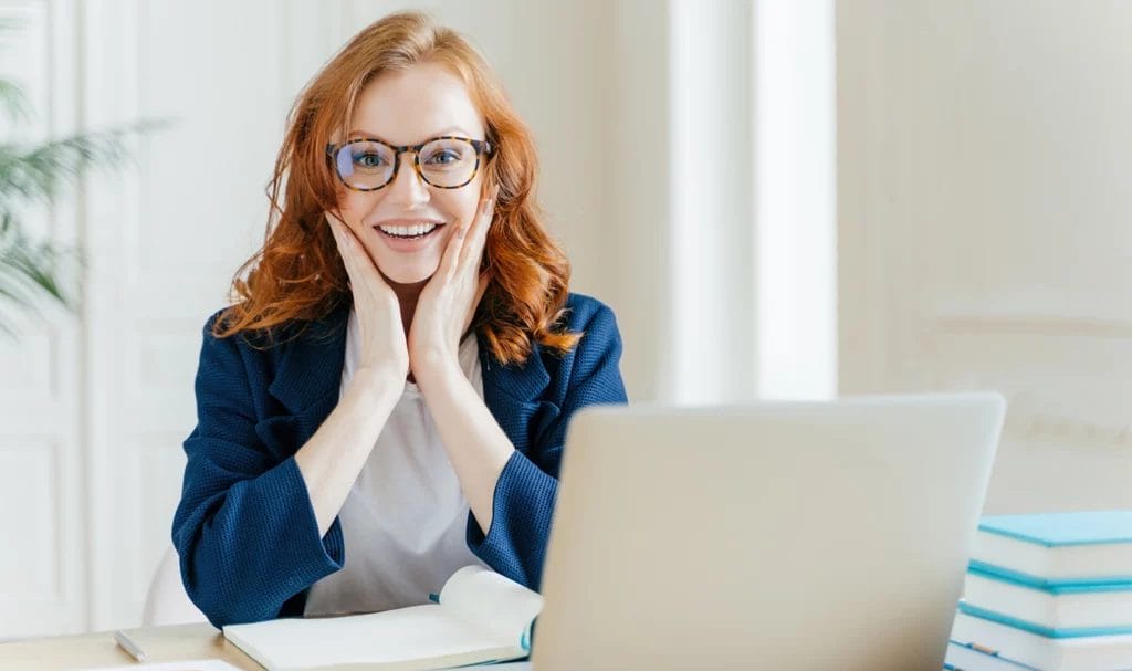Happy consulant has hands on cheeks while smiling gleefully at the camera next to her laptop and notes after a client call.