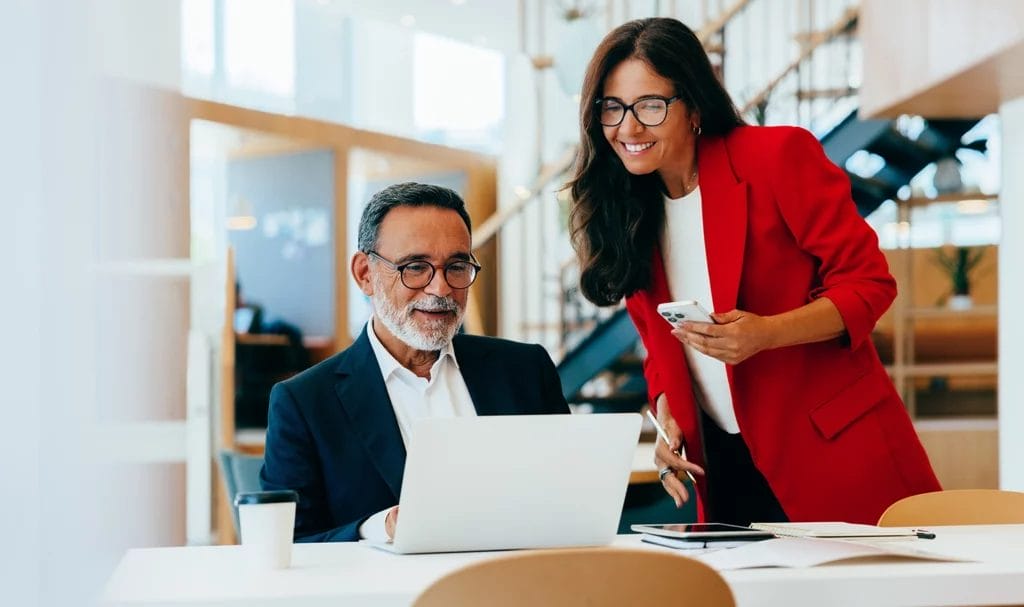 Two senior consultanting professionals working together in an office environment with one on a laptop and the other on a smartphone.