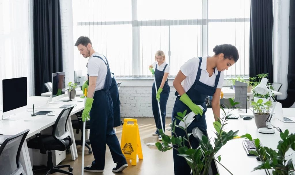 A cleaning crew is mopping and wipping down desks in an open office space next to large windows.