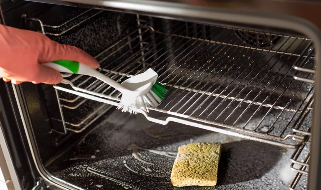 A close up look at a house keeper deep cleaning an oven with a scrubber brush and a sponge.