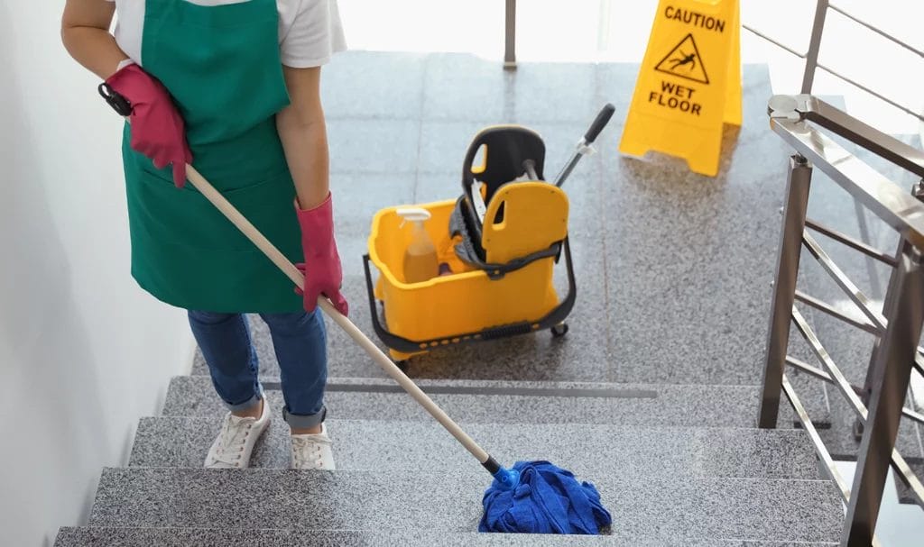 A cleaner is carefully mopping each step of the concrete floors in an office building. A mop bucket and wet floor sign are on the stair landing behind them.