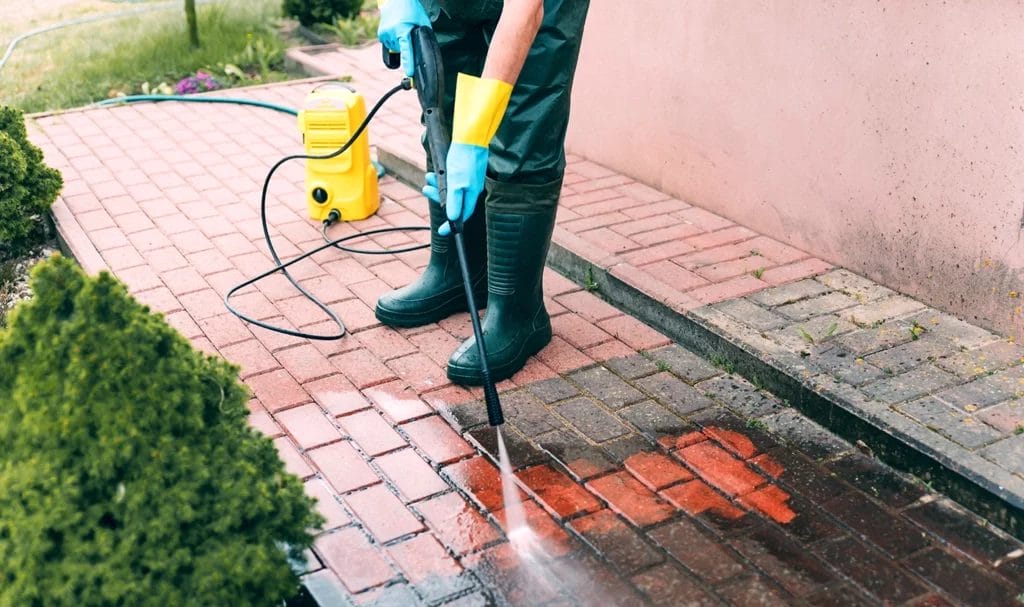 A pressure washer is using a high powered hose to clean dirt and debris off a brick walkway outside of a home.