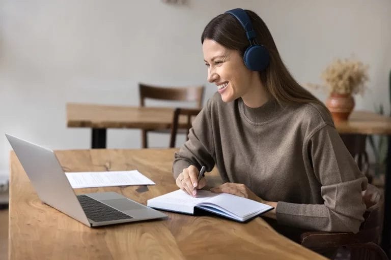 A young woman wears a headset and smiles at her laptop webcam while taking notes during an online course.