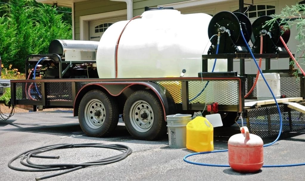 A tank of water, along with other cleaning solutions and hoses, are on a trailer or the front driveway of a home being serviced by pressure washers.