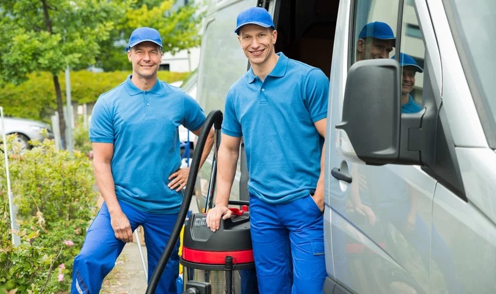 Two pressure washing professionals smile and pose outside of their cleaning van.