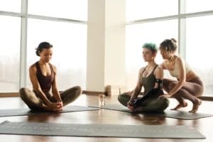Two yoga students sit in lotus position while their instructor adjusts their posture.