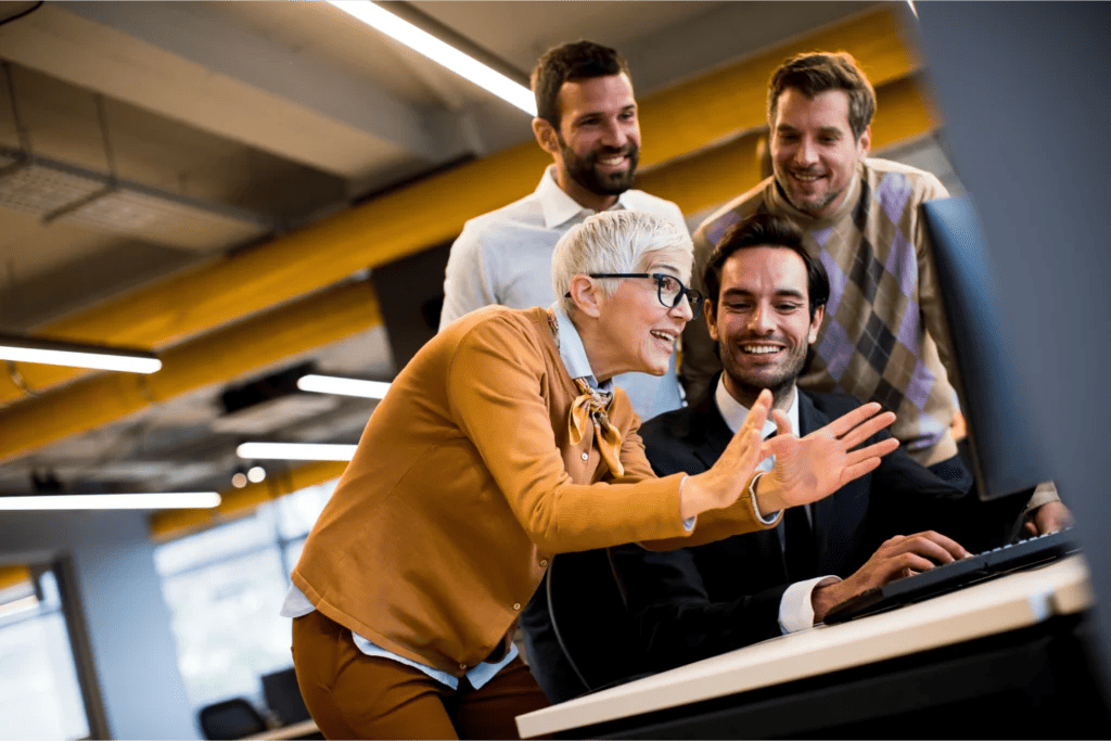 Mature business consultant explains a concept to businessmen while they cluster around a computer.