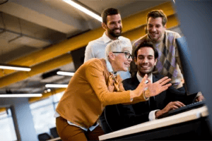 Mature business consultant explains a concept to businessmen while they cluster around a computer.