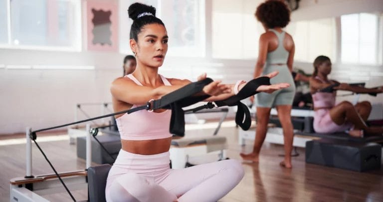 Person in pink athletic wear sitting with legs crossed on a Pilates Reformer machine, performing an exercise.