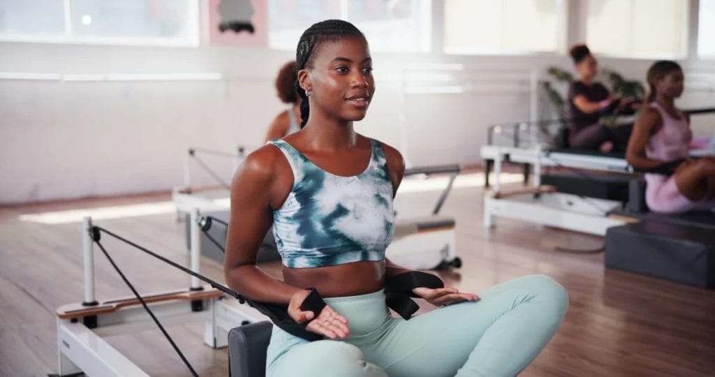 Person in light green and blue athletic wear performs a seated exercise on a Pilates Reformer machine