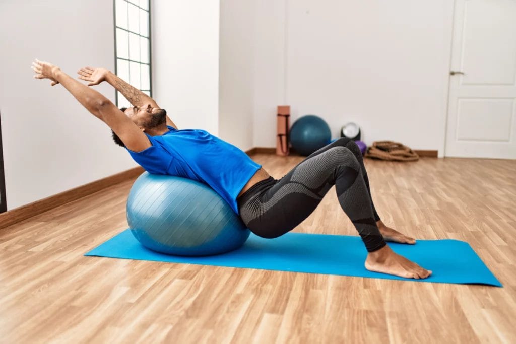 A bearded man in blue performs a Pilates exercise on a mat with a physio ball.