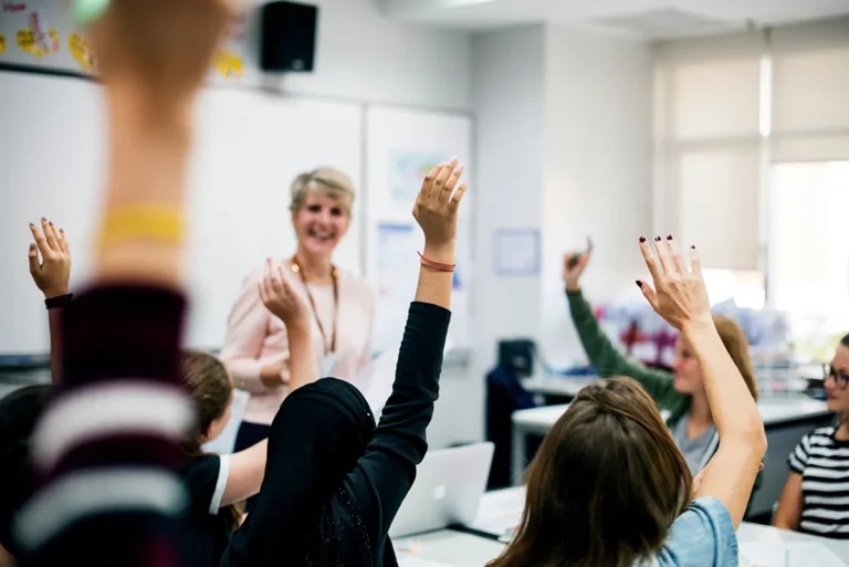 A teacher with short gray hair and a pink cardigan stands in front of a classroom full of students, some of whom have their hands raised.