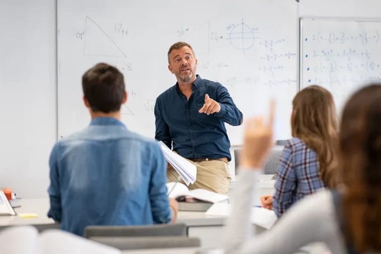 A male teacher stands in front of a whiteboard with mathematical equations written on it and calls on a student in the foreground with her hand raised.