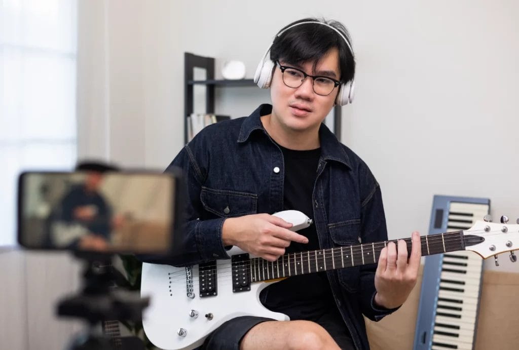 A man wearing a denim jacket and white headphones holds a white electric guitar and films himself with a smartphone on a tripod in an at-home music studio.