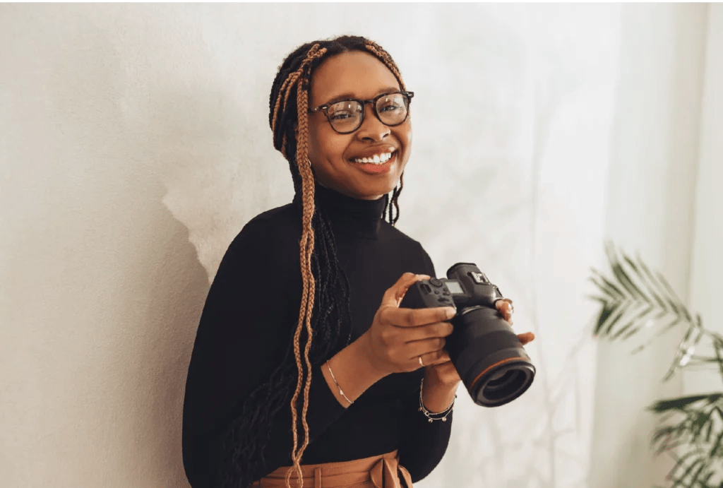 A photographer wearing glasses and a black turtleneck top stands against a white wall, holds a camera, and smiles.